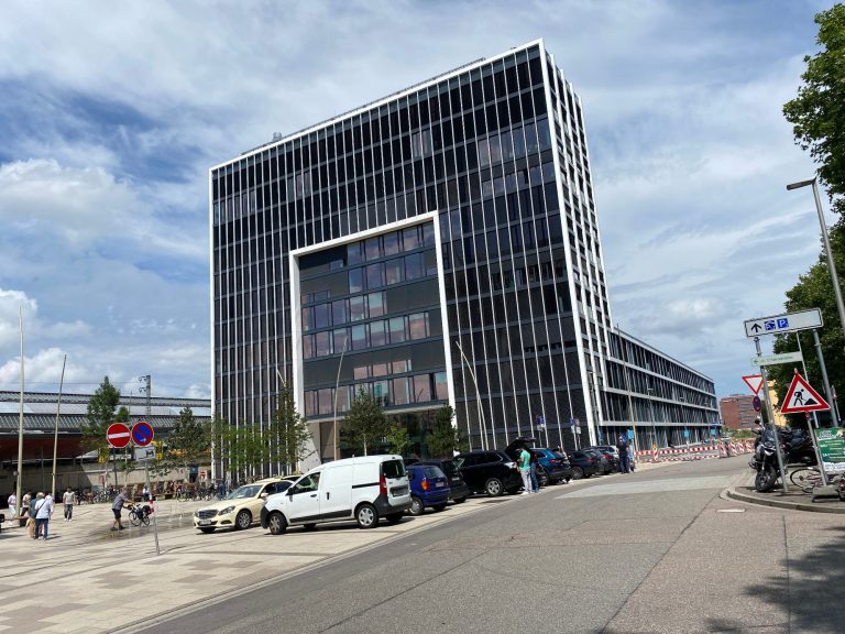 Montage Gutachten durch Sachverständiger Alexander Nestle Moderne Gebäude mit großer Glasfassade, umgeben von Bäumen und Verkehr.